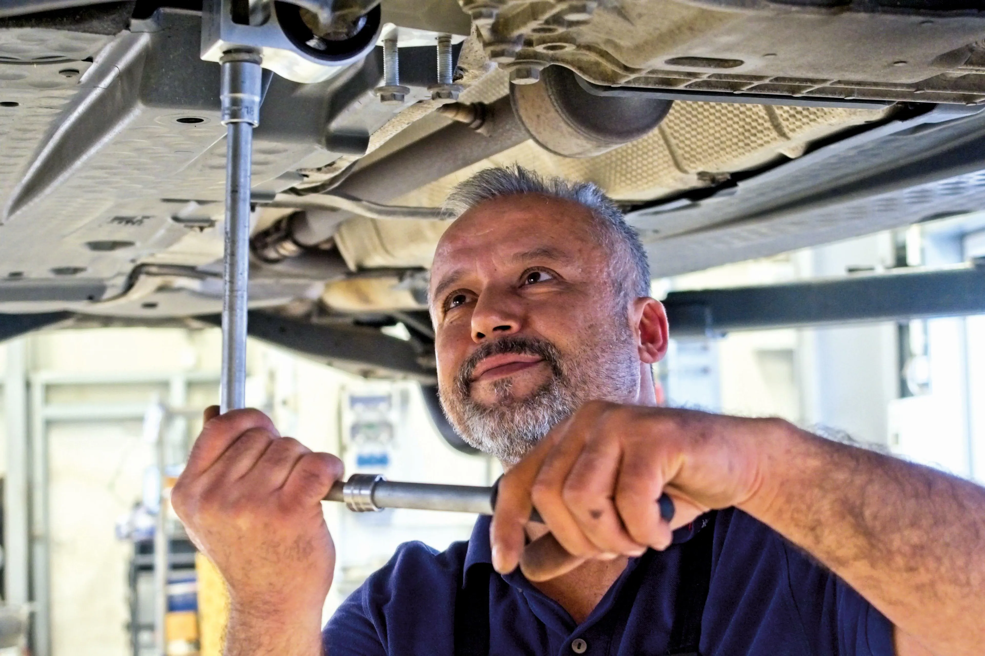 Unsere KFZ-Mechaniker lösen jedes Problem Es wird ein Automechaniker gezeigt, der mit einem Schraubenschlüssel eine Schraube am Unterboden eines Fahrzeugs löst.